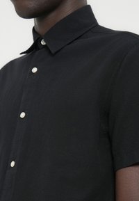 Μαύρο πουκάμισο με κοντά μανίκια από απαλό ύφασμα, με κλασικό γιακά και λευκά κουμπιά. Απλός σχεδιασμός χωρίς σχέδια ή διακοσμήσεις.