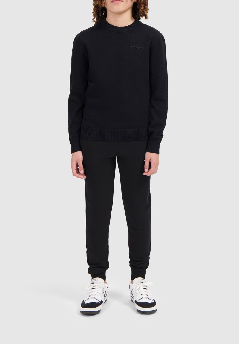 Zwarte gebreide trui met geribde ronde hals en manchetten, gecombineerd met zwarte joggers en witte sneakers met zwarte accenten. Eenvoudig, getailleerd ontwerp.