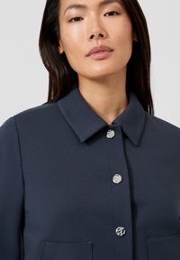 Granatowa koszula z kołnierzykiem, zapięciem na srebrne guziki, teksturowaną tkaniną oraz dwoma przednimi kieszeniami. Prosty, dopasowany krój.