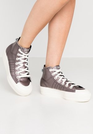 Jambes portant des baskets montantes Adidas gris métallisé avec semelles blanches épaisses et lacets sur un fond clair.