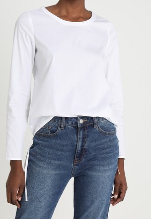Persona que lleva una camiseta blanca lisa de manga larga metida en unos vaqueros azul de talle alto sobre un fondo neutro.