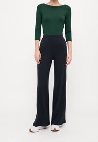 Langsleeve, aansluitend donkergroen topje gecombineerd met hoge, geribbelde zwarte wijde broek. Witte schoenen met beige accenten maken de outfit compleet.