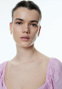 Close-up van een persoon met een lichte huid en bruine ogen, die gouden hangoorbellen en een roze, textuurachtige top met geplooide details draagt.