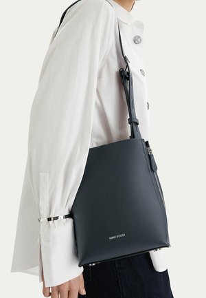 Person wearing white buttoned shirt carries black Gino Rossi leather shoulder bag with zipper and adjustable strap.