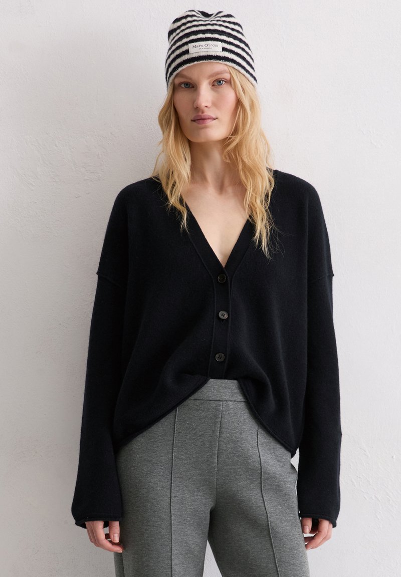 Cardigan noir surdimensionné avec un profond décolleté en V et quatre boutons, associé à un pantalon gris clair ajusté. Bonnet rayé avec étiquette logo.