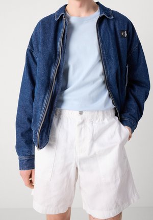 Person trägt eine dunkelblaue Jeansjacke über einem hellblauen T-Shirt und lockere weiße Shorts, Hände teilweise in den Taschen.