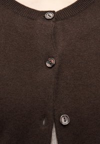 Καφέ πλεκτή ζακέτα με τέσσερα μαύρα κουμπιά κουμπωμένα μπροστά, που αφήνει να φαίνεται ένα μικρό τμήμα του λαιμού και του πάνω μέρους του στήθους.