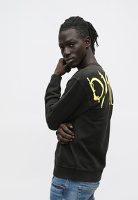 Νεαρός με σκούρες κοτσίδες φορούσε μαύρο φούτερ με κίτρινα γράμματα στην πλάτη και μπλε τζιν, ποζάροντας πλαγίως σε λευκό φόντο.