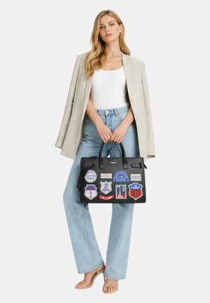 Femme portant un blazer beige, un top blanc, un jean bleu clair, des talons nude, tenant un sac à main noir avec des patchs colorés sur le thème de Paris.