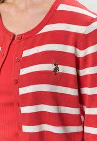 Červený kardigán s bielymi horizontálnymi pruhmi, s okrúhlym výstrihom a zapínaním na gombíky. Vyšívané logo na ľavej strane. Mäkka textura.