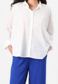 Biała koszula zapinana na guziki o luźnym kroju, z dekoracyjnymi, teksturowanymi akcentami w kształcie pereł, w połączeniu z niebieskimi spodniami o szerokich nogawkach.