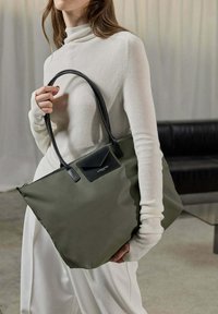 Borsa tote verde oliva con una forma strutturata, dettagli in pelle nera e chiusura con zip. Modello che indossa una maglietta bianca a costine e pantaloni chiari.