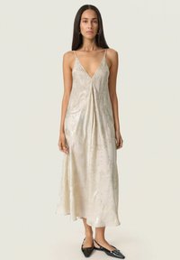 Woman with long dark hair wearing a sleeveless, shiny cream midi dress with thin straps and black pointed flat shoes.
