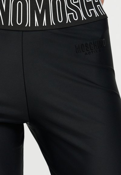Κοντινό πλάνο μαύρων κολάν Moschino Active με την εκτύπωση «Moschino» στη μέση σε λευκό χρώμα και με ανάγλυφο λογότυπο στον μηρό.