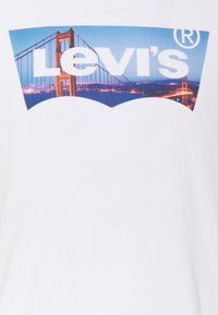 Valkoinen puuvillapaita, jossa on graafinen kuva Golden Gate -sillasta ja "Levi's"-logo paksuilla kirjaimilla keskellä.