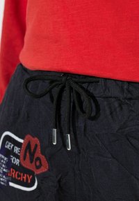 Schwarze Shorts mit Kordelzug und strukturierter Oberfläche, versehen mit einem roten und blauen grafischen Patch. Kombiniert mit einem roten Langarmshirt.