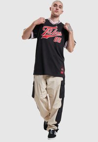 Zwarte sporttrui met rode letters, gecombineerd met beige cargobroek met zwarte zijkanten en meerdere zakken. Zwarte sneakers.