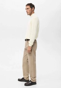 Suéter color crema, pantalones beige y zapatos oscuros. El conjunto presenta un diseño ajustado con puños acanalados. Texturas suaves y una paleta de colores neutros.