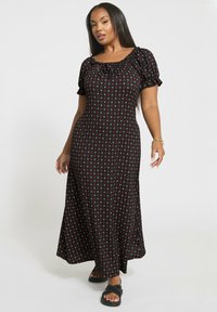 Kvinna i en svart maxiklänning med små röda och vita blommönster, korta puffärmar och svarta sandaler, stående mot en vit bakgrund.