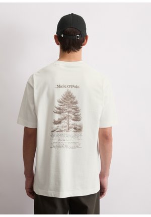 Mężczyzna w białej koszulce Marc O'Polo z detaliczną ilustracją sosny i opisowym tekstem na plecach, czarna czapka, neutralne tło.