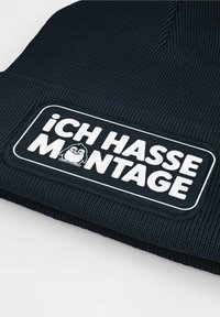Black ribbed knit beanie featuring a rectangular white patch with the text "ICH HASSE MONTAGE" and a cartoon penguin graphic.