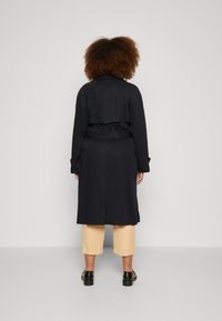 Marineblå trenchcoat med belte i midjen, lange ermer og lagdelt bakdetalj. Brukt over lysebeige trekvartbukser og svarte sko.