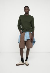 Pulover verde tricotat, pantaloni scurți cargo maro cu buzunare, cămașă din denim albastru deschis legată în talie, adidași albi, șosete negre.