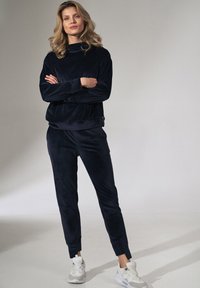 Marineblauwe velours trainingspak met een hoge hals en taps toelopende broek. Glad textuur, elastische manchetten en zijzakken. Draagt met lichte sneakers.