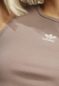 Brązowy, ribbedowy top sportowy z okrągłym dekoltem, z białym haftowanym logo Adidas na lewej piersi.
