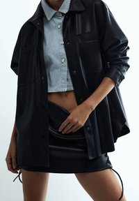 Black leather oversized shirt jacket layered over a cropped striped blue shirt, paired with a black mini skirt featuring drawstring sides.