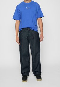 Blå bomullströja med en vit logotyp, tillsammans med mörka jeans i lös passform och svarta sneakers med ljusbruna sulor.