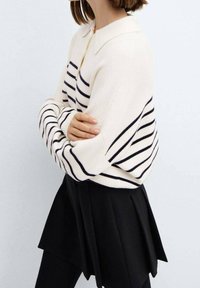 Pull crème avec des rayures horizontales noires, doté d'un col et de manches oversized, associé à une jupe plissée noire.