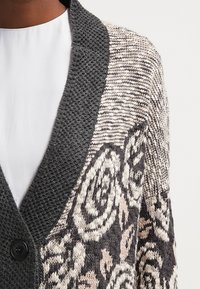 Sweter kardigan z dzianiny, z ciemnoszarym teksturowanym kołnierzem i kremowym wzorem róż na korpusie, zapinany z przodu na guziki.