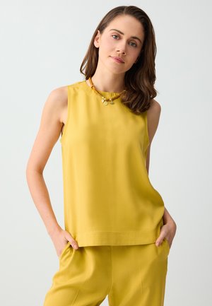 Jeune femme aux cheveux bruns mi-longs, portant un top jaune sans manches et un pantalon jaune assorti, mains dans les poches, fond neutre.