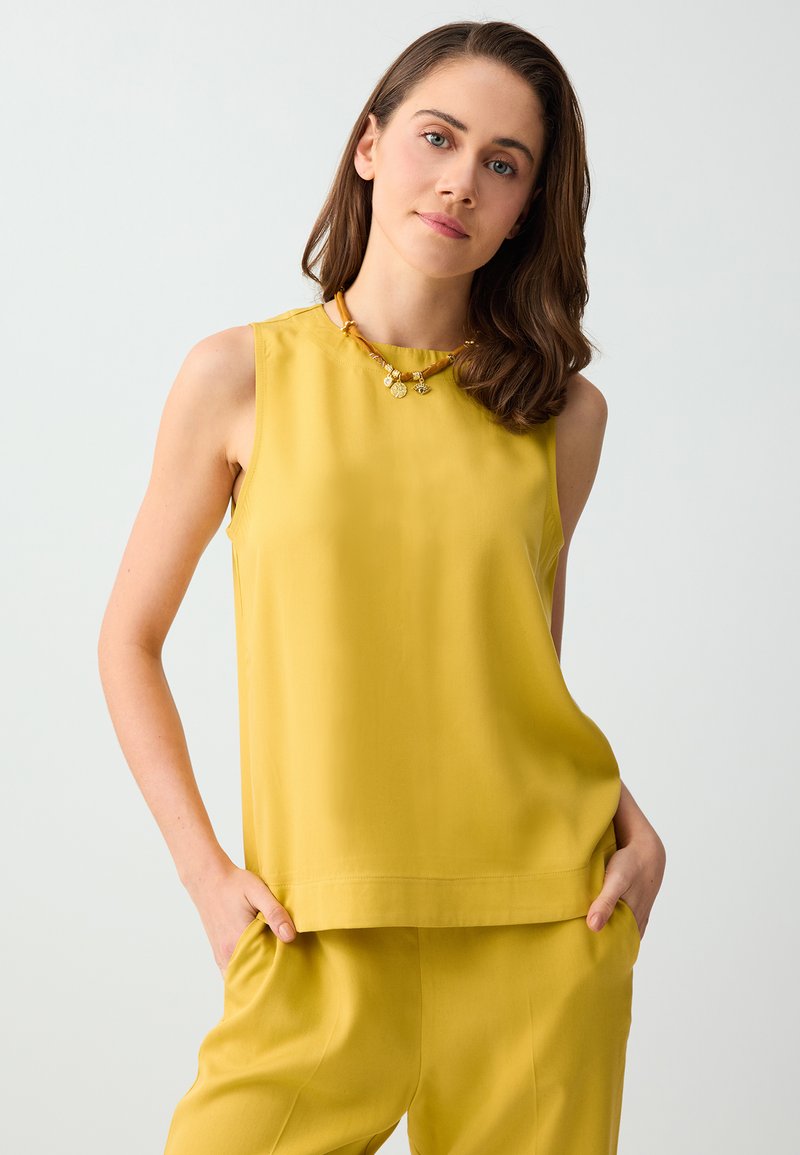 Jeune femme aux cheveux bruns mi-longs, portant un top jaune sans manches et un pantalon jaune assorti, mains dans les poches, fond neutre.