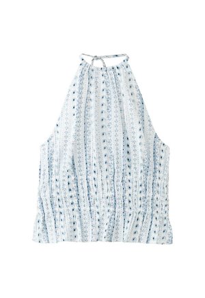 Jeune femme aux cheveux foncés attachés en queue de cheval, portant un haut sans manches à motifs avec col halter, un pantalon blanc à jambes larges et des sandales bleues, debout contre un mur blanc.