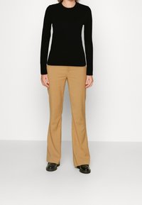 Personne portant un haut à manches longues noir ajusté, un pantalon beige droit et des chaussures noires brillantes, debout devant un fond blanc uni.