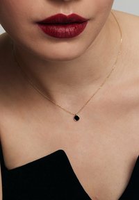 Złoty naszyjnik z łańcuchem i małym czarnym okrągłym wisiorkiem, noszony na odsłoniętej skórze, z widoczną czarną odzieżą z jednej strony.