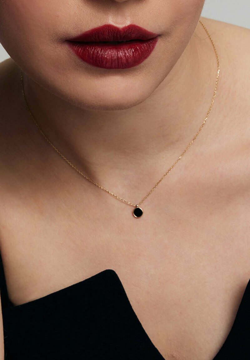 Złoty naszyjnik z łańcuchem i małym czarnym okrągłym wisiorkiem, noszony na odsłoniętej skórze, z widoczną czarną odzieżą z jednej strony.