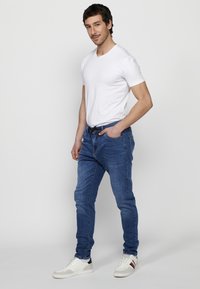 Vaqueros de denim azul con corte recto, llevados con una camiseta blanca de manga corta y zapatillas blancas. El fondo es blanco liso.