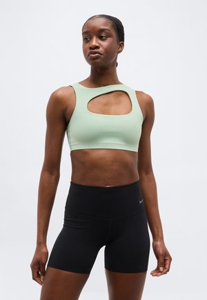 Atletisk kvinde iført en lysegrøn sports-bh med udskæring på brystet og sorte cykelshorts med høj talje, stående mod en ensfarvet baggrund.