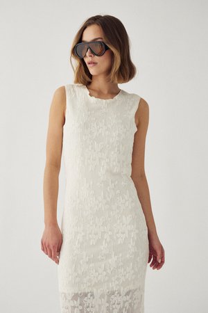 Naine, kes kannab valget varrukateta tekstuurse kleiti ja suuri musti päikeseprille, poseerib ühevärvilise heleda tausta ees.