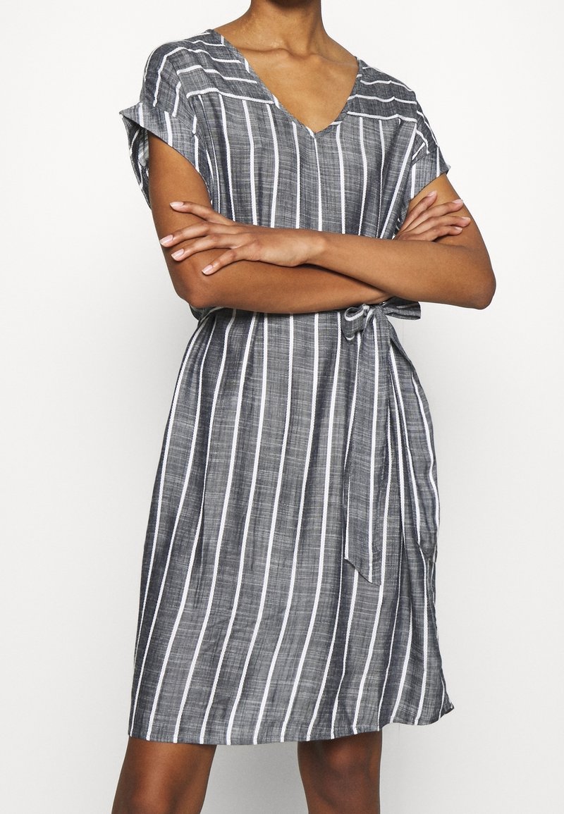 Femme portant une robe grise mi-longue à rayures verticales blanches avec une ceinture à la taille, les bras croisés sur la poitrine.