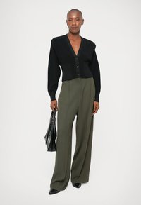 Czarny krótki kardigan z dekoltem w serek i zapięciem na guziki, zestawiony z oliwkowymi spodniami o szerokich nogawkach i czarną torebką.
