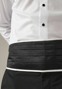 Sort taljeforklæde med plisseret design og hvidt trim, båret over en hvid skjorte med sorte knapper. Stoffet ser glat og holdbart ud.