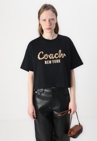 Juoda trumpa marškinėlių su ryškiu "Coach" logotipu gyvūnų raštu, derinama su aukšto juosmens juodomis odos kelnėmis ir mažyte ruda rankine.