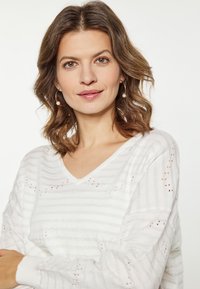 Maglione bianco a V con strisce argento e piccoli dettagli a taglio. Realizzato in morbido tessuto testurizzato. Maniche lunghe con polsini a coste.