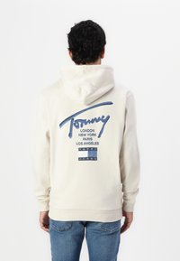 Bež hoodie s kapuljačom i vezicom, koji ima plavi logo i imena gradova na leđima. Mekana tkanina s rebrastim manžetama i donjim rubom.