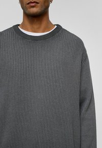 Pull en maille grise avec une texture côtelée, un col rond et des manches longues. Le tissu semble doux et a une coupe décontractée.