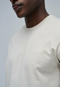 Camiseta de manga corta beige hecha de algodón con cuello redondo. Presenta un sutil detalle de logo en relieve en el pecho. Textura suave.
